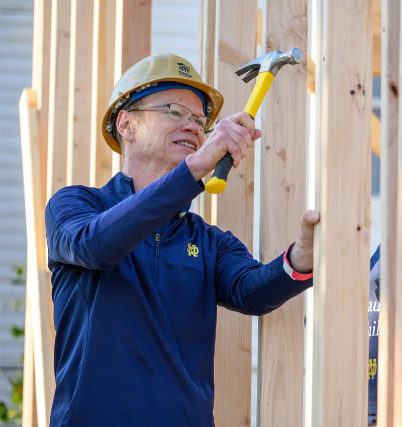 A white man wearing a gold hard hat and a navy blue University of Notre Dame quarter-zip shirt hammers a nail into a wooden frame during a construction project.