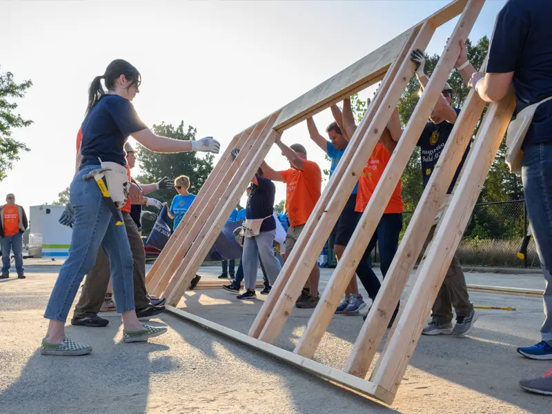 A group of volunteers in orange shirts carefully raise a large wooden wall frame at a community building project. A woman in the foreground guides the frame into place.