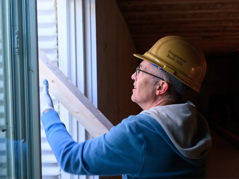 A white man wearing a gold hard hat and work gloves inserts a wooden beam into a window frame during a construction project.