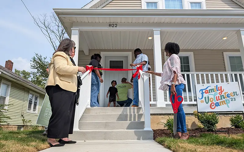 A group of people cut a red ribbon at the entrance of a newly renovated two-story home with the house number 822 visible. A small child stands in the doorway, and a sign on the lawn reads Family Volunteer Camp ‘23.