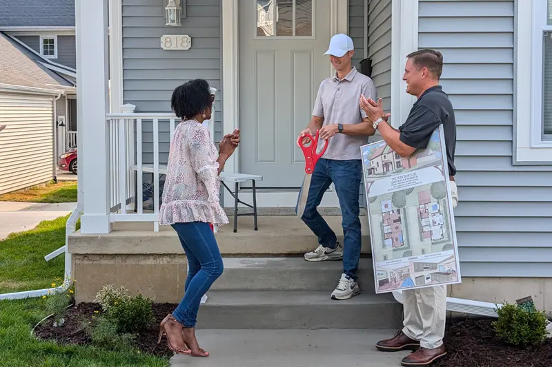 A woman claps and smiles as a man holds a giant red-handled pair of scissors for a ribbon cutting. Another man holds a large rendering of the house's floor plan.