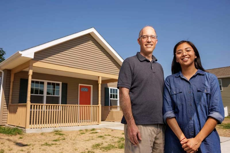 A male professor and female student pose for a photo in front of a one-story home with a bright orange door.