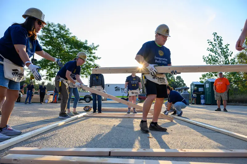 Volunteers wearing navy blue shirts and aprons work together to construct a wooden frame on a paved surface. The sun shines brightly, backlighting some of the volunteers. A Habitat for Humanity box truck is visible in the background.