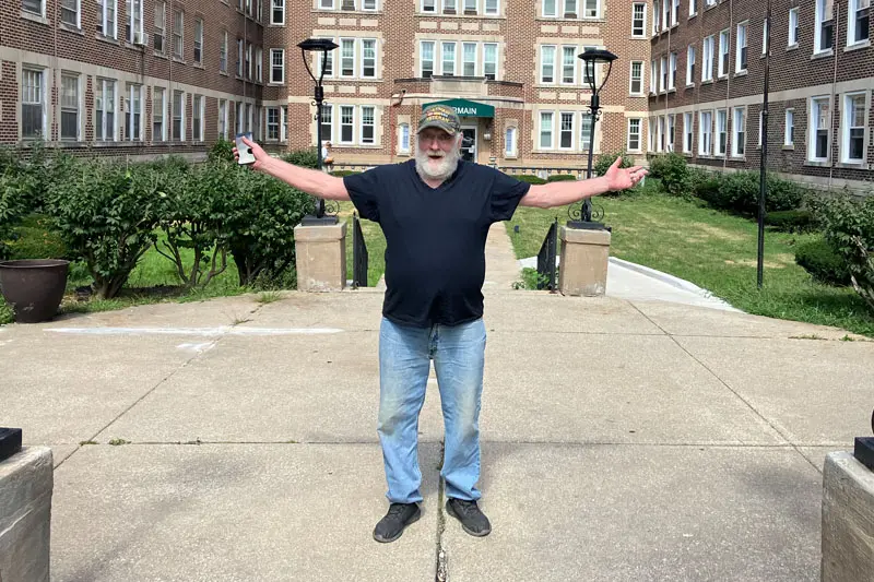 Jack West, a white man, with a white beard stands in front of a brown-bricked apartment building with his arms wide open.