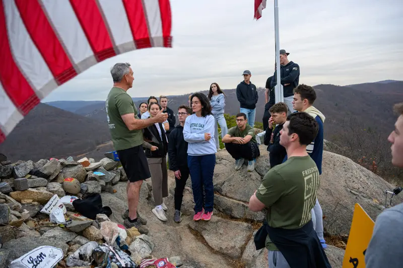 A group of students listens to a speaker atop a rocky summit. A corner of an American flag is visible in the foreground. The students wear a mix of casual clothing and Notre Dame apparel. The speaker, in an olive green shirt, gestures as he addresses them. A weathered flagpole stands behind the group with mountains visible in the background. A small rock cairn sits in the foreground left, adorned with painted stones and small signs.