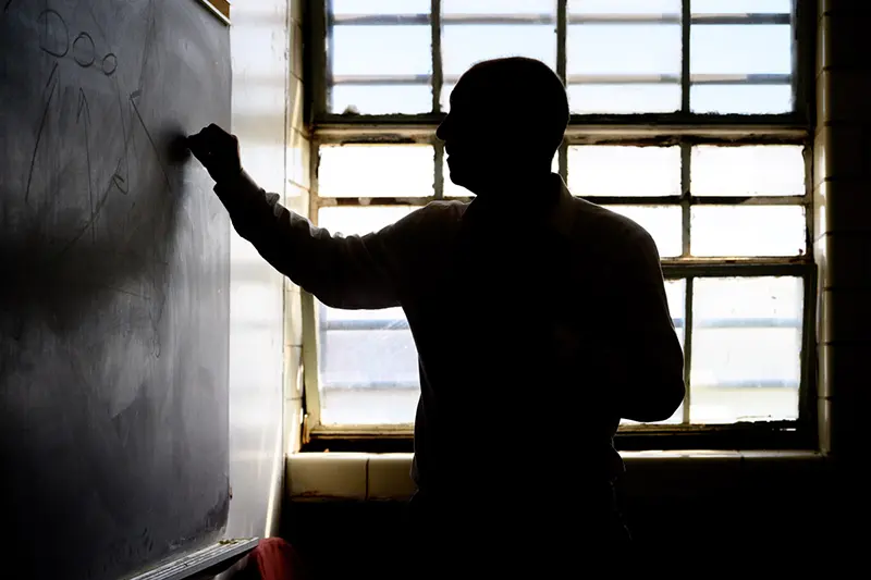 A man stands in profile writing on a chalkboard. He is silhouetted against a bright window.