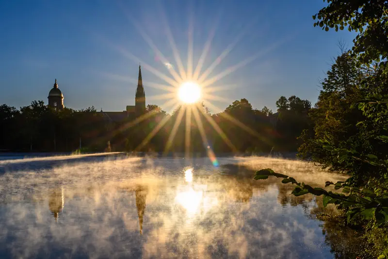 The sun rises over St. Mary's Lake on the University of Notre Dame campus. The Golden Dome and Basilica of the Sacred Heart are silhouetted against the brilliant sunburst. Wisps of fog drift across the lake, reflecting the light and the iconic landmarks. Green trees frame the right side of the image.