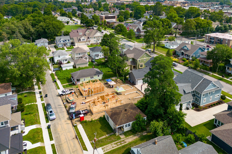 Aerial view of a residential neighborhood with two homes under construction. The wooden frames of the houses are visible, and construction workers are on site. Trucks and equipment are parked nearby. Surrounding the construction site are established homes with yards and driveways.