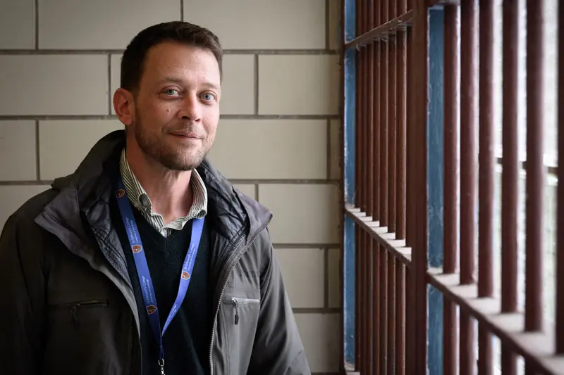 A white man wearing a gray winter jacket and a lanyard is standing next to prison bars.