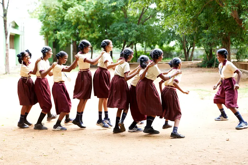 A line of Indiana girls in matching tan and maroon school uniforms hold each other's shoulders and playfully skip across a dirt schoolyard. Trees and a school building can be seen in the background.