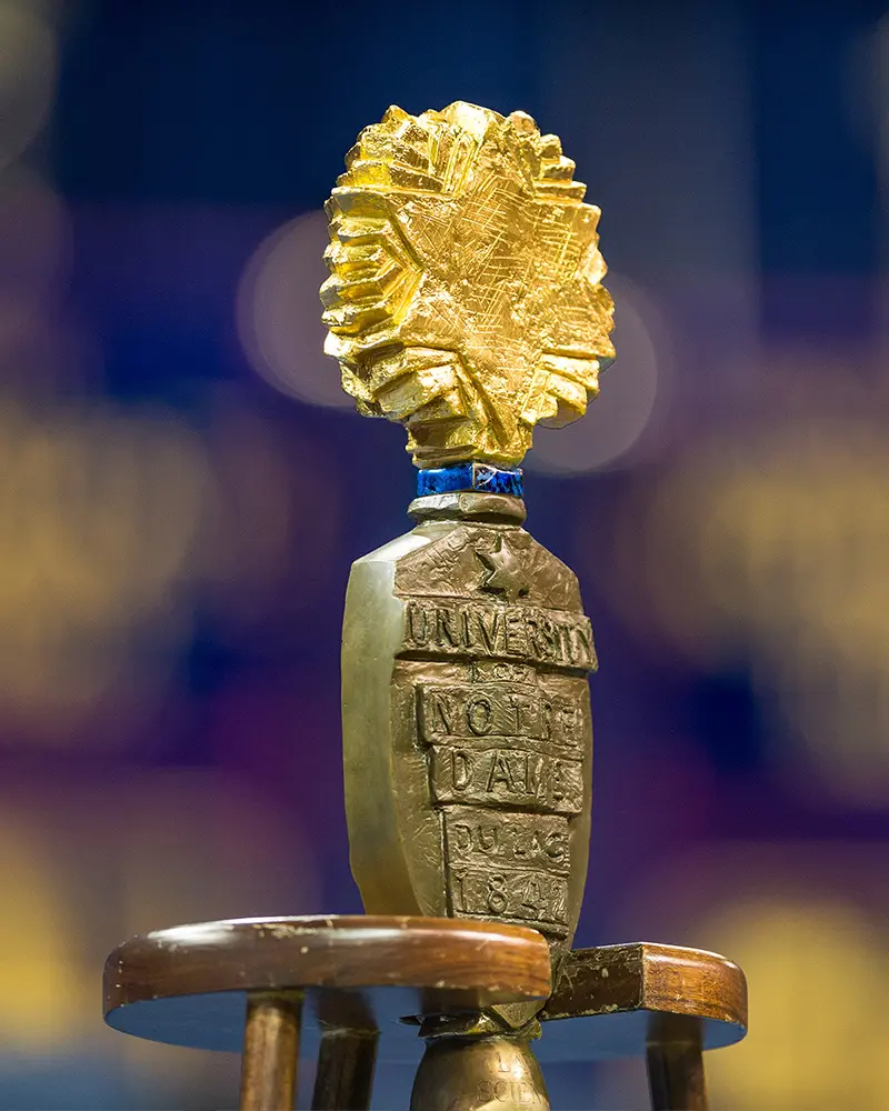 A close up of the University Mace features a bronze shield inscribed with University of Notre Dame du Lac 1842 below a golden burst of stars