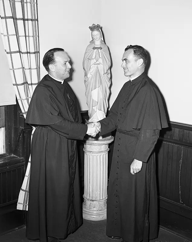 A black and white image of Fr. Cavanaugh and Fr. Hesburgh shaking hands in front of a statue of Mary. 