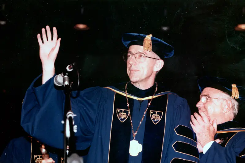 Newly inaugurated President Malloy stands in his academic robe at a podium, waving at the crowd.