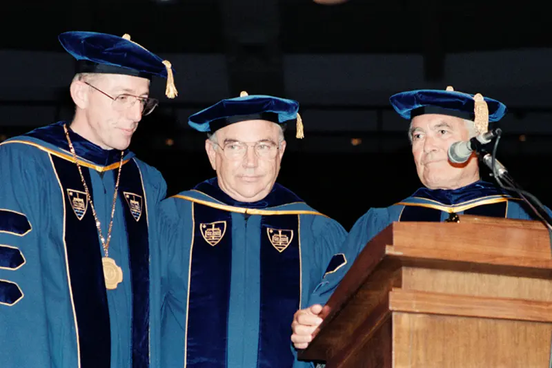 Rev. Edward Malloy, C.S.C., Donald Keough, and Rev. Theodore M. Hesburgh, C.S.C. stand at a podium in their academic robes.