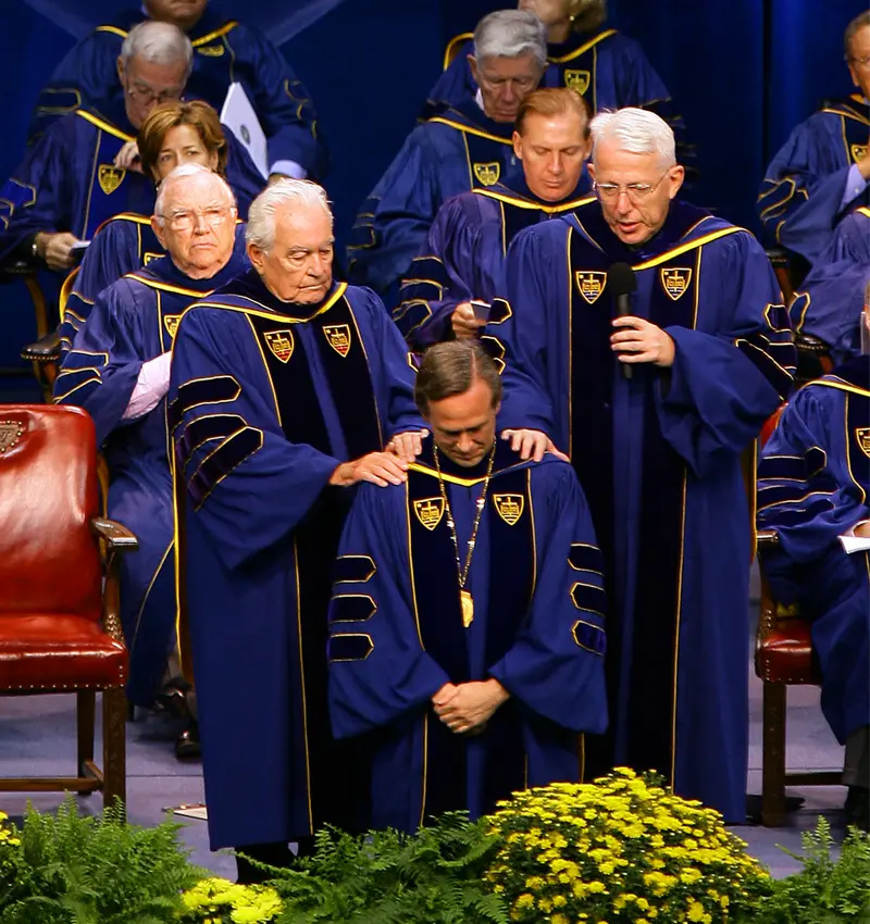 Fr. Jenkins kneels on stage with Fr. Hesburgh and Fr. Malloy lay their hands on him in blessing.