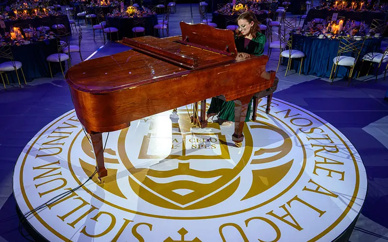 Miriam Rice sits at a piano on a stage in the middle of a room full of empty round tables.