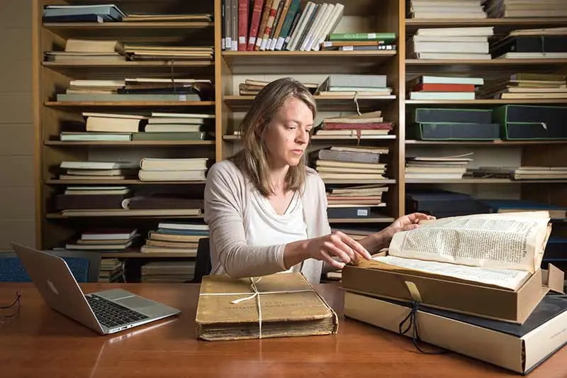 Elaine Stratton Hild at her desk, flips through an old book. Bookshelves line the wall behind her and her laptop is open on her desk