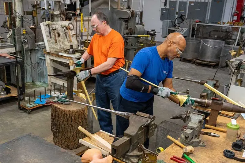 A photo of two men, one White and one Black, shape bells by hand using hammers and mandrels.