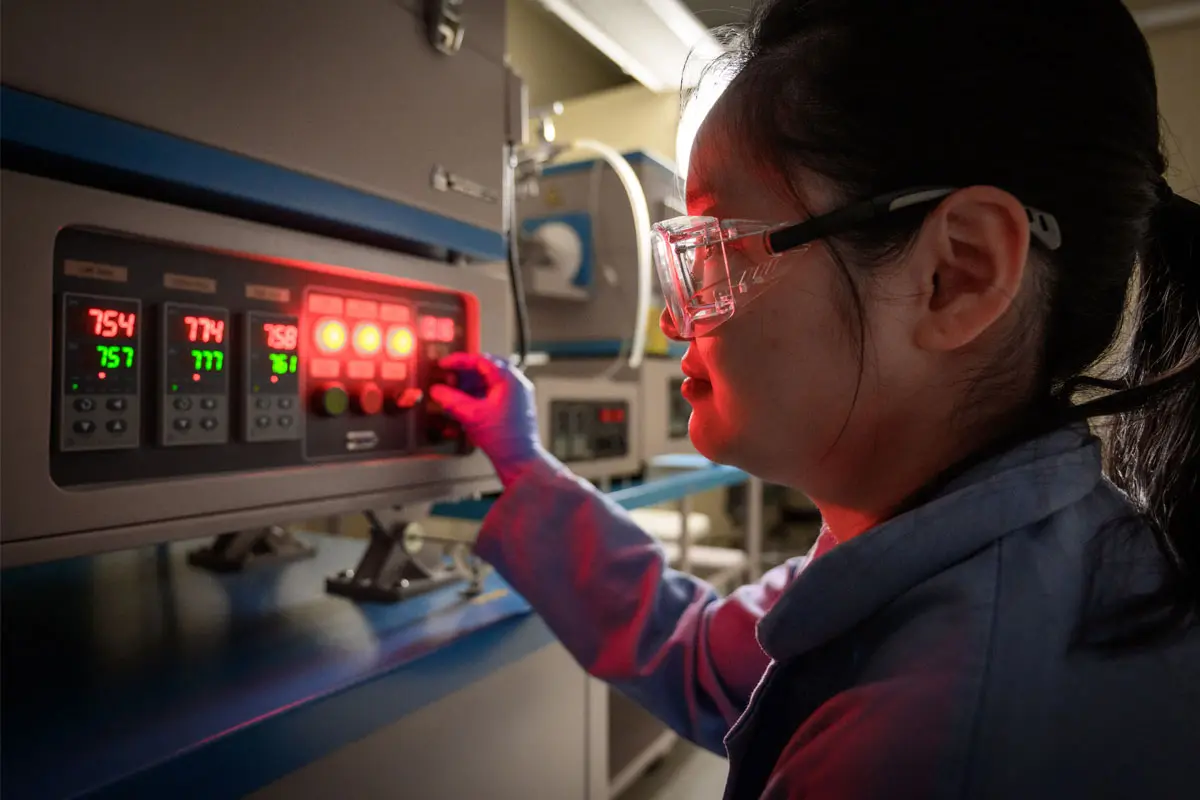 A female student wearing safety goggles adjusts knobs on machinery.