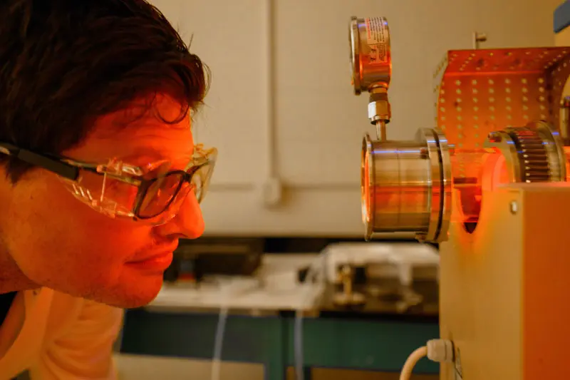 A man wearing safety goggles over glass looks closely at an incinerator.