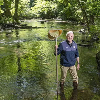 Gary Lamberti standing in a creek with waders and a net taking samples from the creek bed.