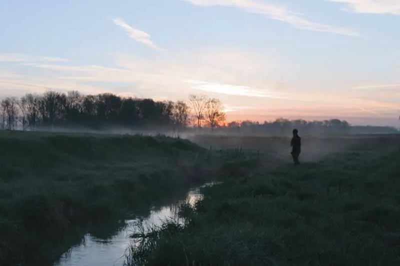 A person standing by a small river in the fog of the early morning.