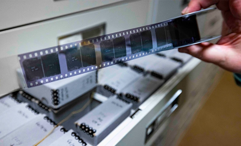 A hand holds a strip of developed film over an open drawer filled with archival boxes. The film contains a series of dark, exposed frames.