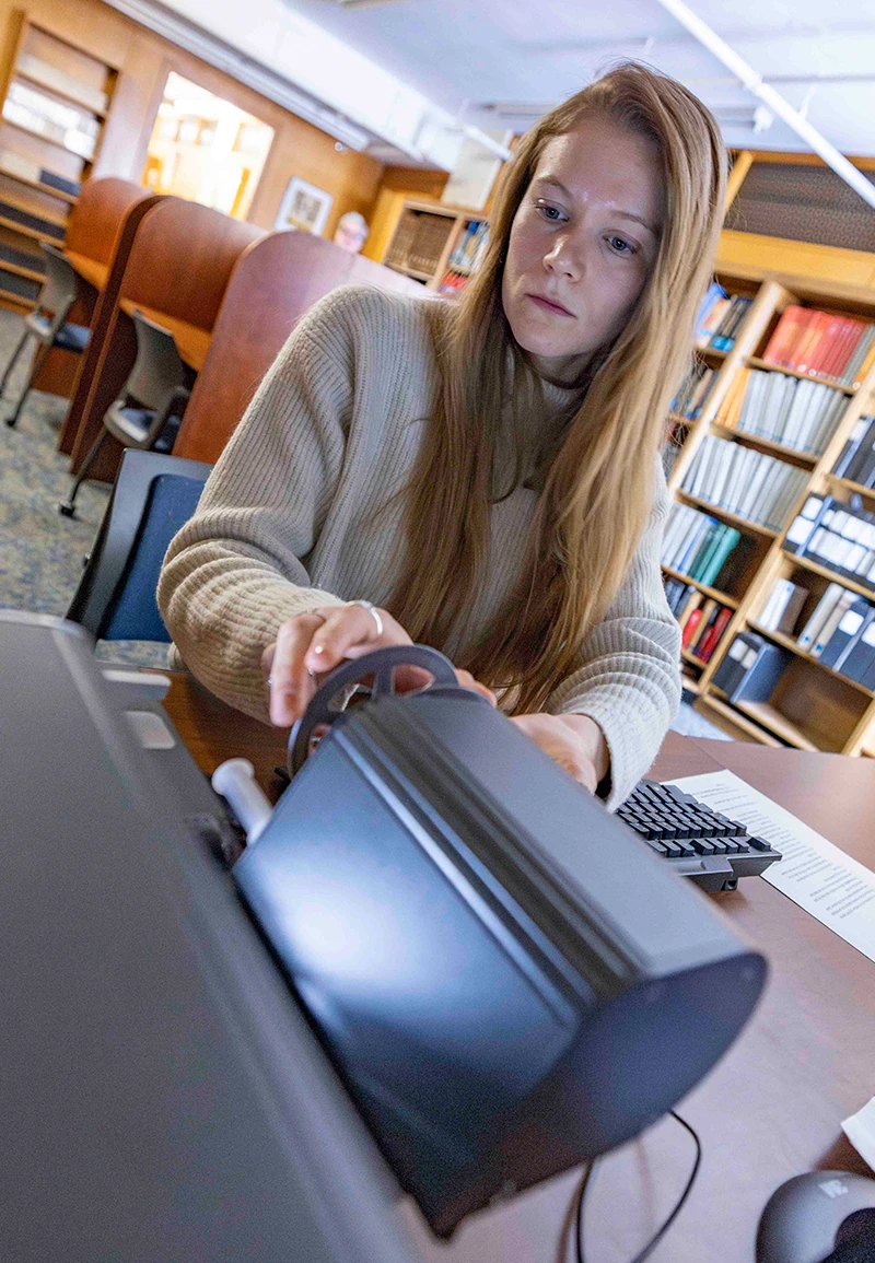 A student with long, light brown hair uses a microfilm reader in a library. Wooden bookshelves line the wall behind her.