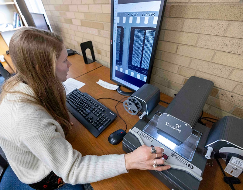 A student digitizes microfilm using a STImaging machine. The monitor displays a magnified view of the microfilm text, and the student carefully operates the machine to capture the images.