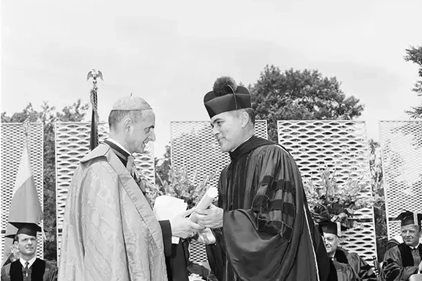 Black and white photo of a graduation ceremony. A person in academic robes and biretta receives a diploma from a cardinal. Other graduates and faculty are seated in the background. Large ferns decorate the stage.