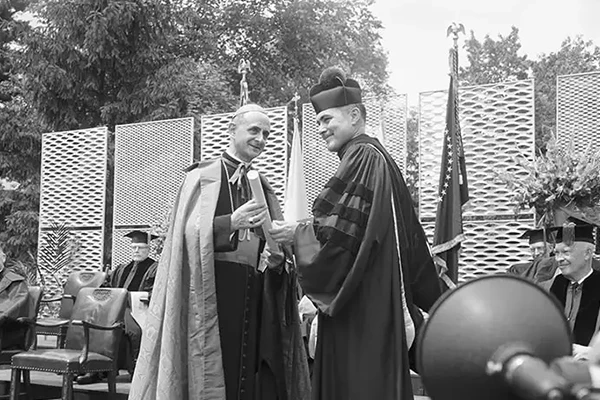 A black and white photo of a graduation ceremony. A person in academic robes receives a diploma from a clergyman in a bishop's mitre and cope. Other figures in academic dress are seated behind them on a stage. A decorative screen and trees form the backdrop. A large microphone is partially visible in the foreground.