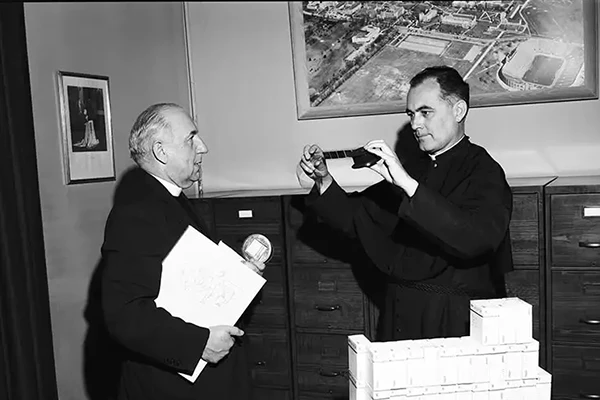 Black and white photo of two priests in an office. One priest holds a book and a small, circular award. The other priest examines microfilm. A framed aerial photo of Notre Dame's campus hangs on the wall. A large stack of white boxes sits on a table.