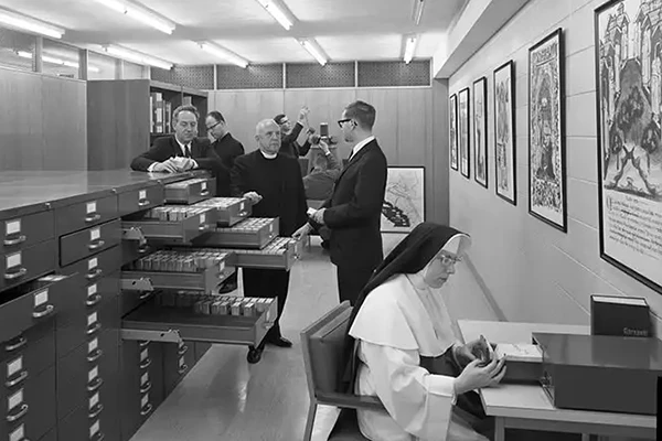 Black and white photo of the University Archives. Several men in suits, including a priest, examine open drawers of a large filing cabinet. Framed artwork hangs on the wall, and a nun sits at a desk in the foreground, working with archival materials. Fluorescent lights illuminate the room.