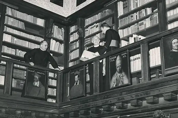 Black and white photo of a corner in a multi-level library. Three priests stand on the upper level amidst bookshelves. Framed portraits of religious figures hang on the railing.