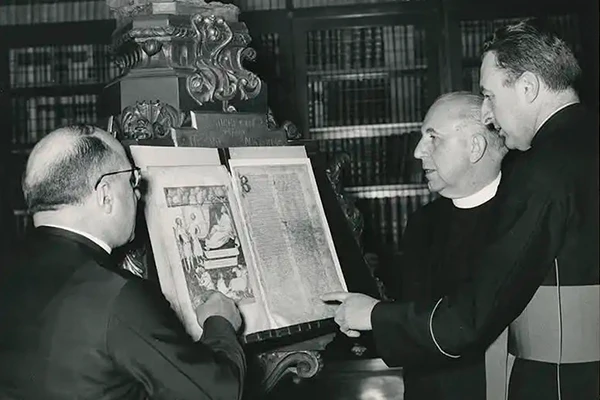 Black and white photo of three priests examining an open book displayed on a stand. The book features illustrations on one side and text on the other. The priests are dressed in clerical garb and appear to be discussing the book's contents in a library setting. A decorative statue is visible in the background, indicating they may be in an archive or special collections room.