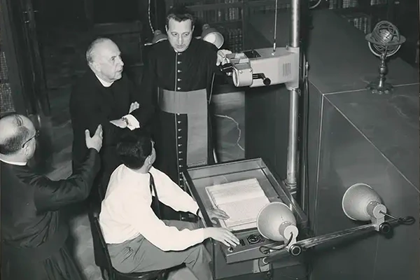 Black and white photo of four men and a microfilm reader. One man sits at the reader, with an open book positioned beneath its lens. Two men in clerical garb stand behind him, observing. Another man in clerical garb gestures towards the machine. A small globe sits atop a cabinet in the background.