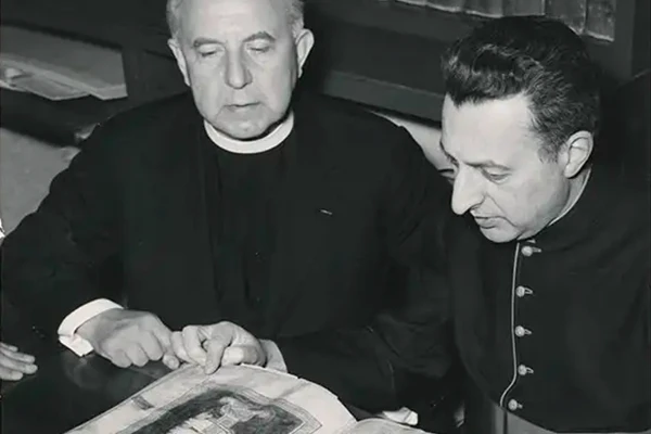 Black and white photo of two men examining a large, open book. The book's pages display intricate illustrations, possibly of religious or historical scenes. One man, dressed in clerical garb, looks down at the book. The other man, wearing a dark jacket with buttons, points to a detail on the page. Bookshelves filled with volumes line the background.