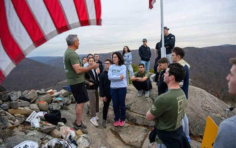 A group of people stand at the top of the Trail of the Fallen near West Point, a steep hike that ends at a large mound of stones that have been carried up and placed in memory of soldiers who have died in military conflicts. In the foreground is the American flag.