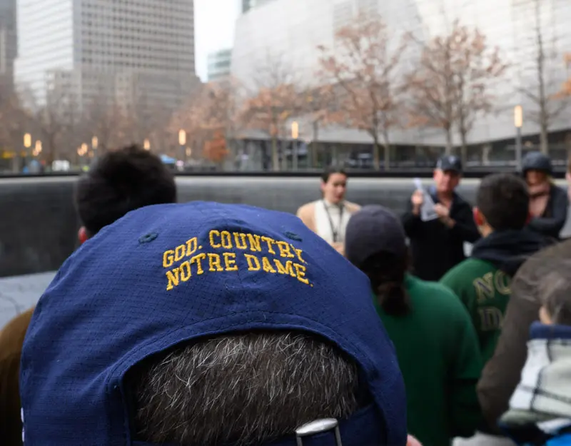 A close up of the back of a navy hat with the words God, Country, Notre Dame on it. The person is standing at the nine eleven memorial listening to a presenter.