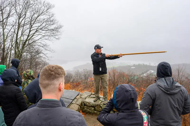 A white man stands at the top of the Trail of the Fallen near West Point, talking to a group of cadets. He's pointing his walking stick at something out in the distance.