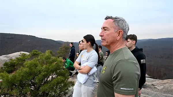 A white man stands at the top of a mountain talking military strategy to a group of cadets.