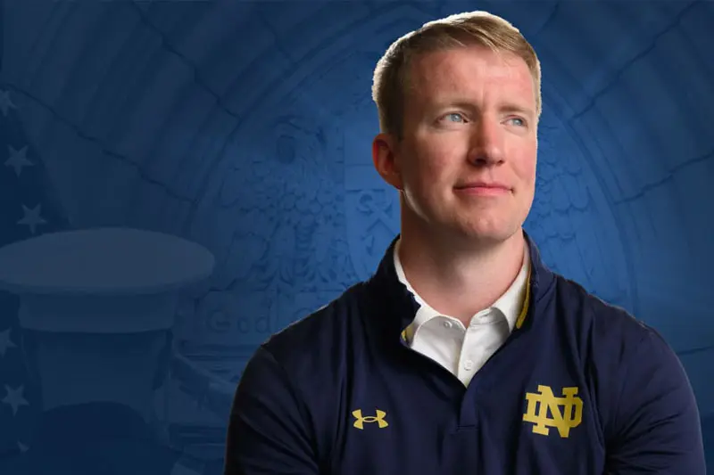 A white man in a navy blue Under Armour quarter-zip pullover with the gold Notre Dame logo looks to the upper right.