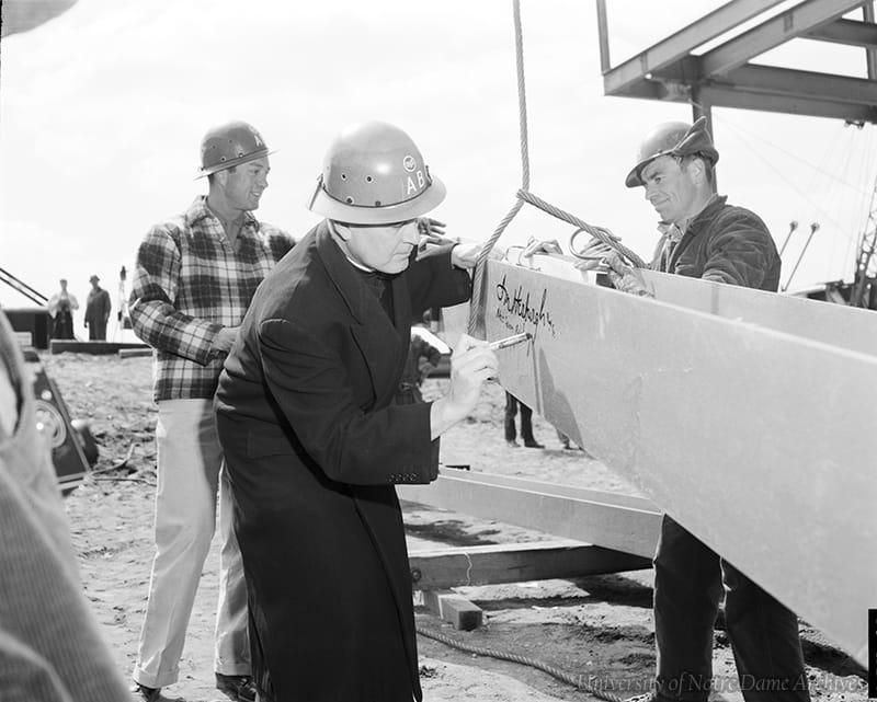 Father Hesburgh, wearing a hard hat, signs a steel beam, while construction works look on.