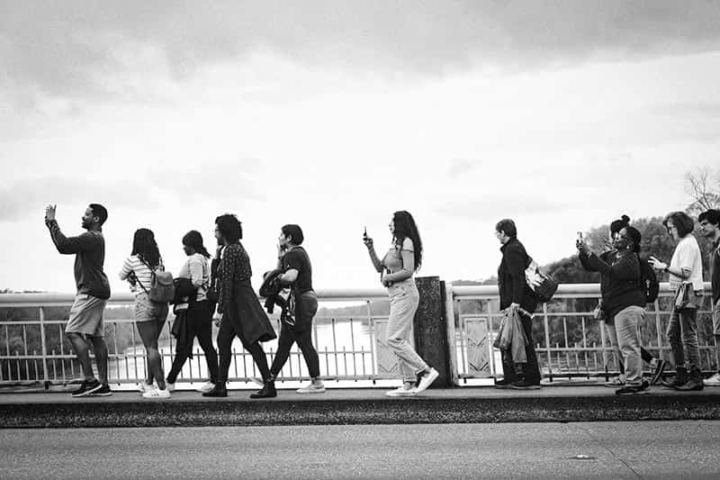 Students walk across the Edmund Pettus Bridge with their phones out taking video and photos.