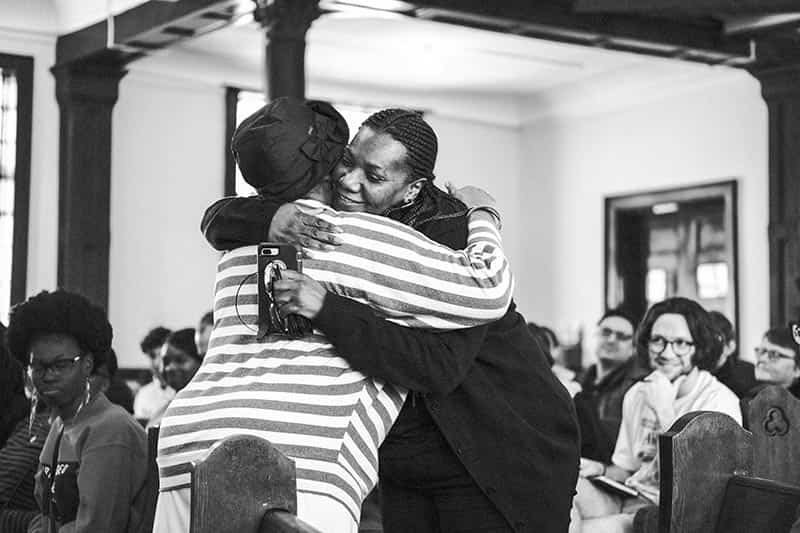 Two women hug in the middle of an aisle of a church.