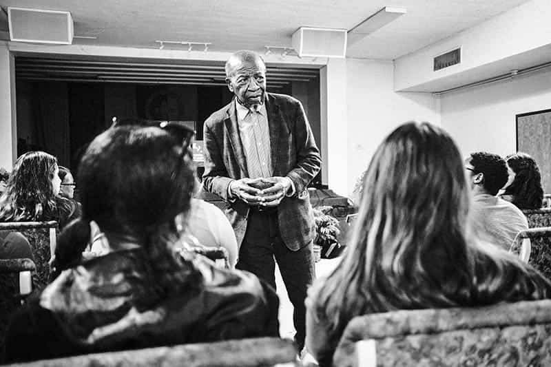 A man holds his hands together, standing in front of students.