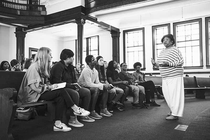 A woman talks to students sitting at church pews.