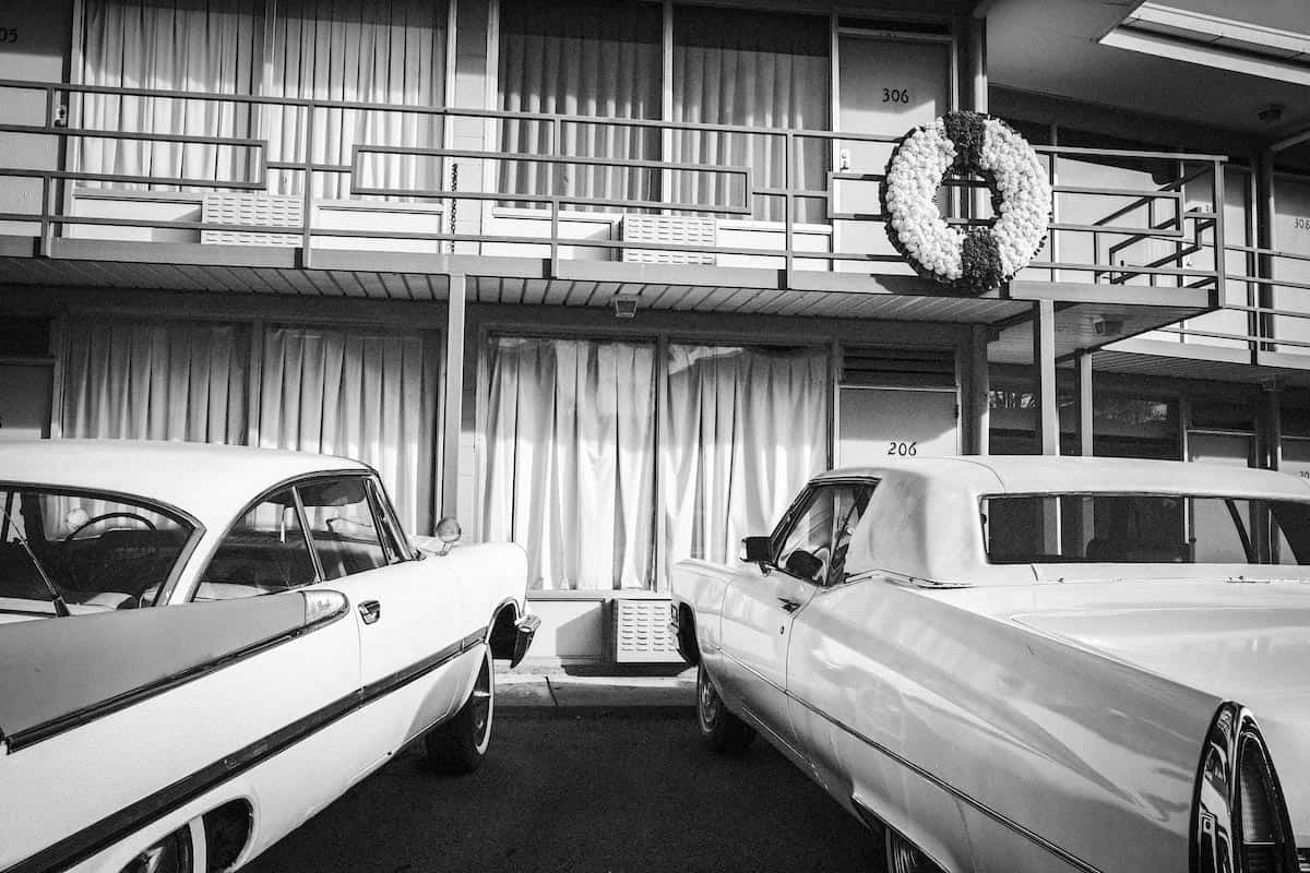 Two cars parked outside of a hotel balcony, a wreath hangs outside one of the rooms.