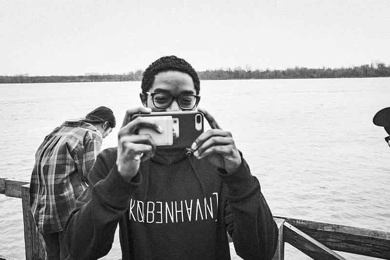 A student pointing his phone camera at the the photographer, a river is in the background.