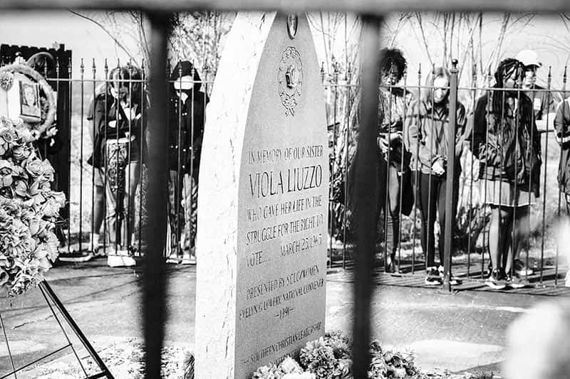 People stand around a grave that is blocked by a tall, iron fence.